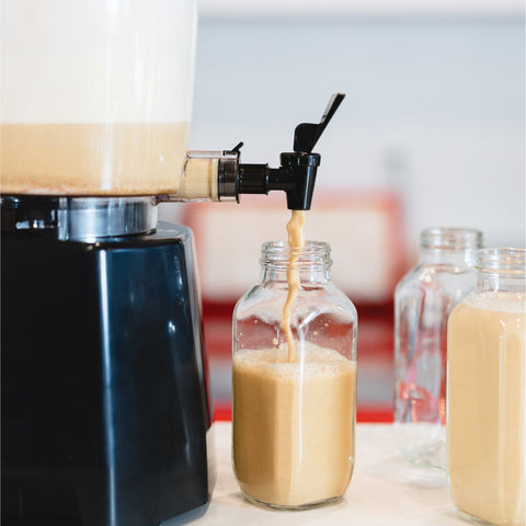 A Nutramilk dispenses a brown nut milk into square glass bottles.